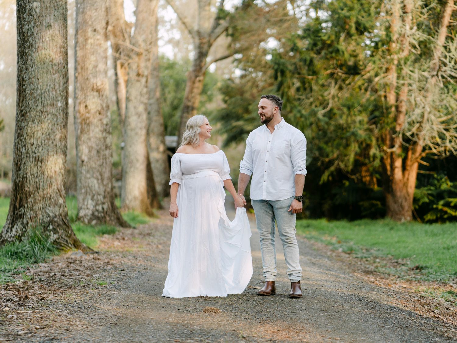Couple walking hand in hand on a tree-lined dirt path, woman in white dress, man in white shirt and khakis.