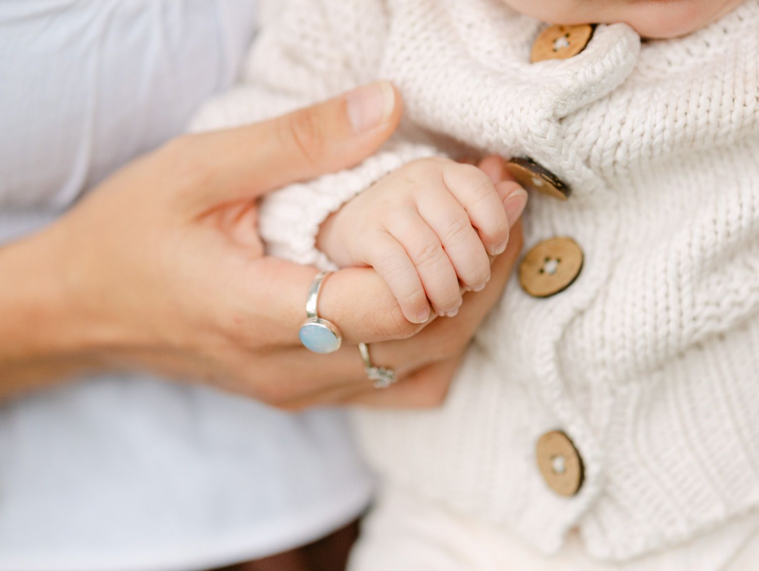 Close-up of a newborn baby holding an adult's hand, wearing a knit sweater with buttons.