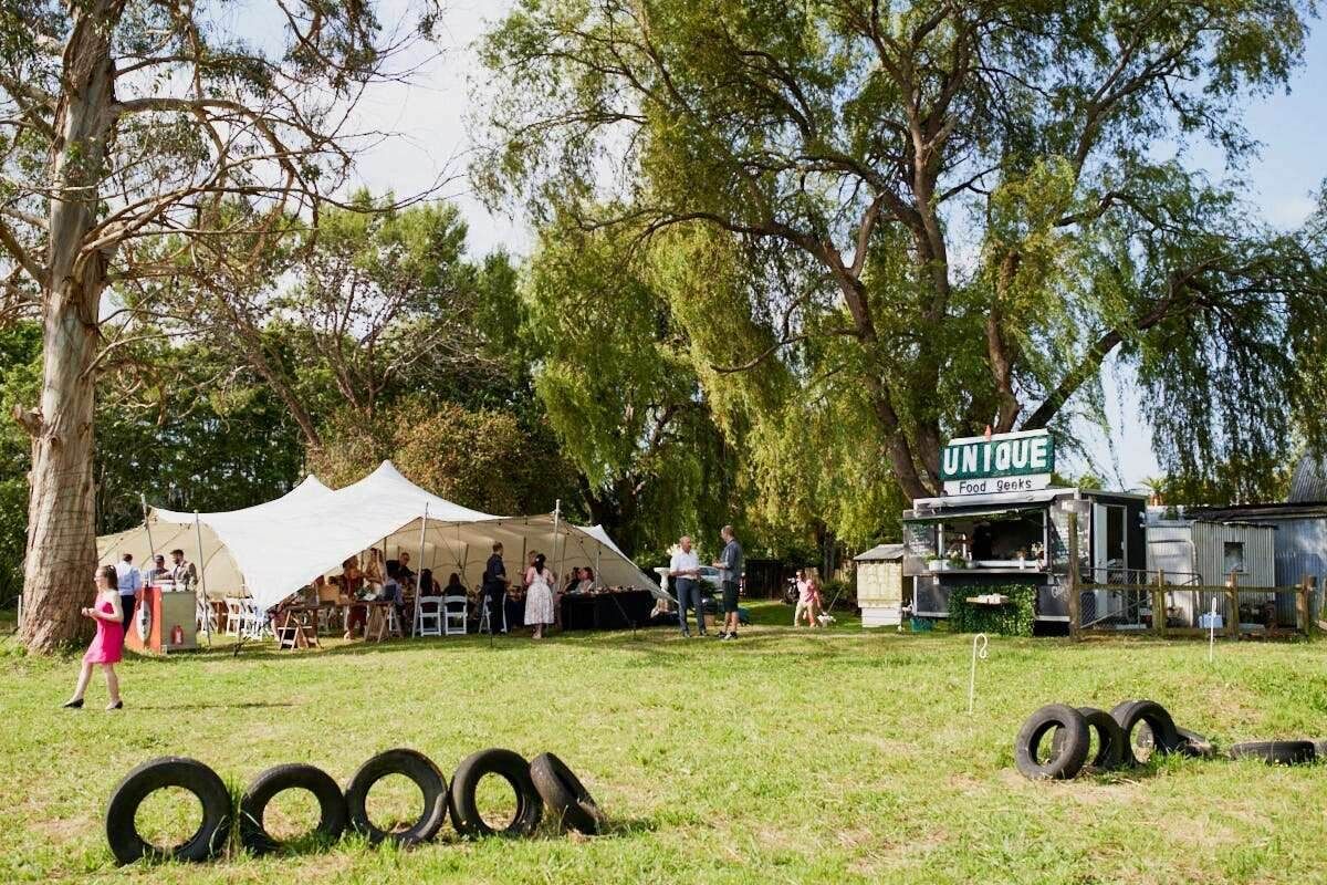 Outdoor festival with white tents, food truck, tire decorations, and people gathered under large trees on a sunny day.