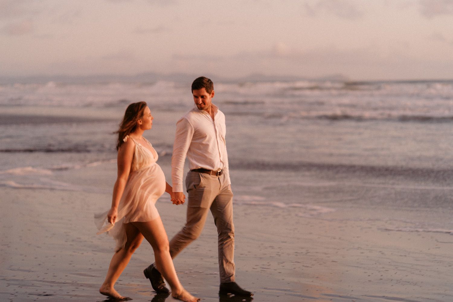 Pregnant couple walking hand in hand on a beach at sunset, wearing white outfits with waves in the background.