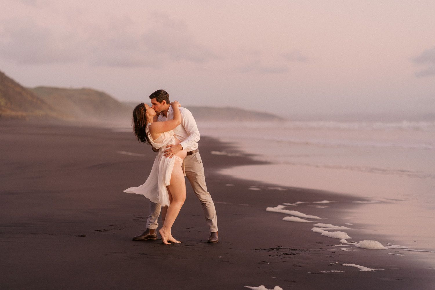 Couple sharing a romantic kiss on a black sand beach at sunset, waves and misty hills in the background.