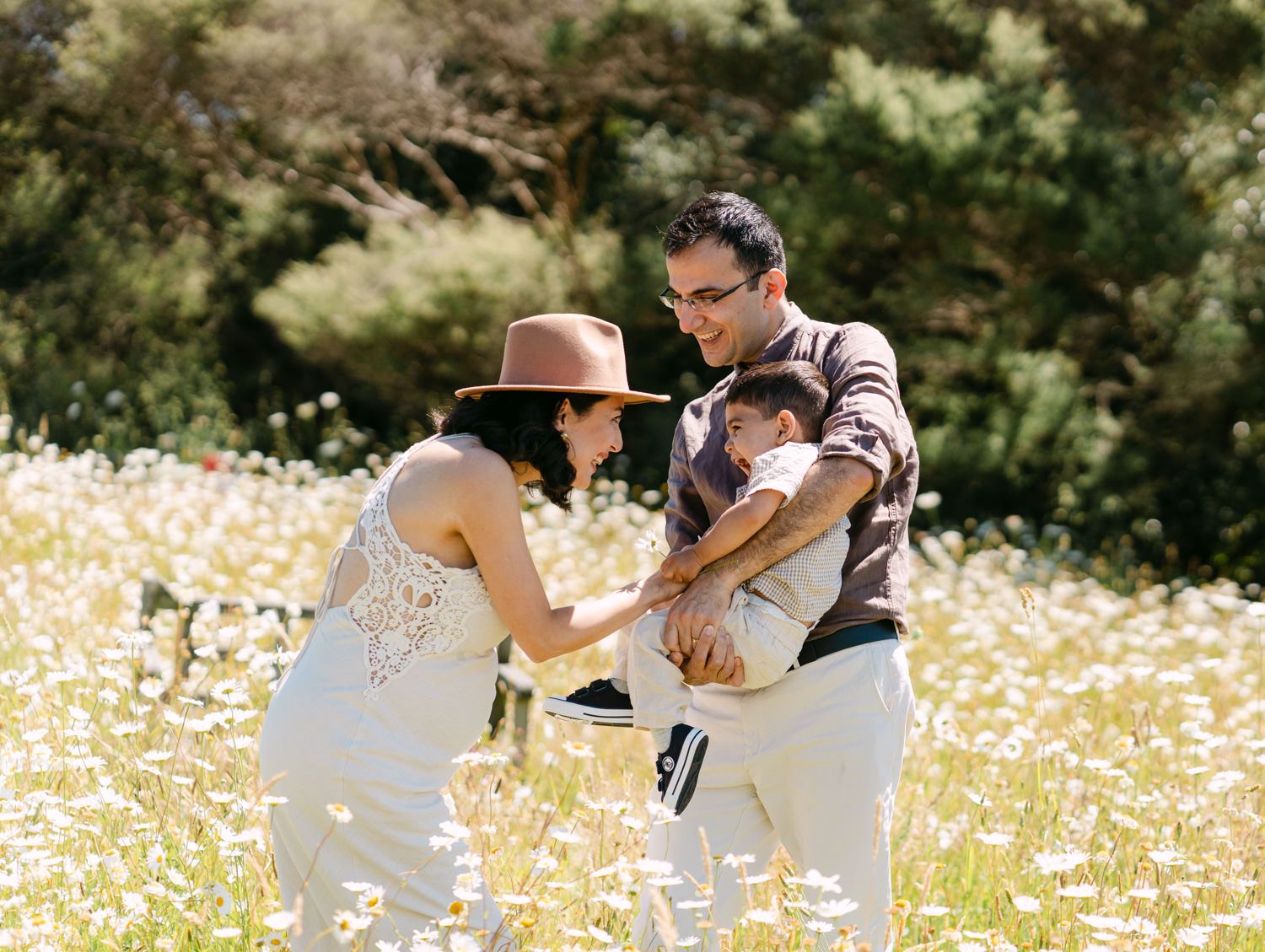 Family playing in a sunny daisy field, parents laughing with child, lush green trees in background.
