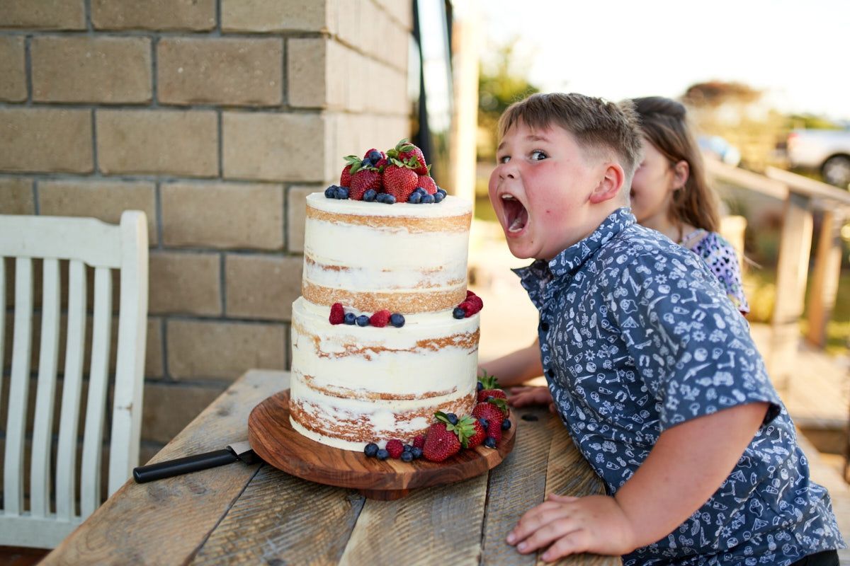 Boy excitedly eyeing a two-tier naked wedding cake topped with fresh strawberries and berries on a rustic wooden table.