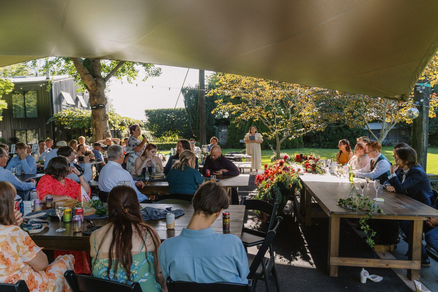 Guests gathered under a canopy at an outdoor garden party, seated at long tables surrounded by lush green trees.