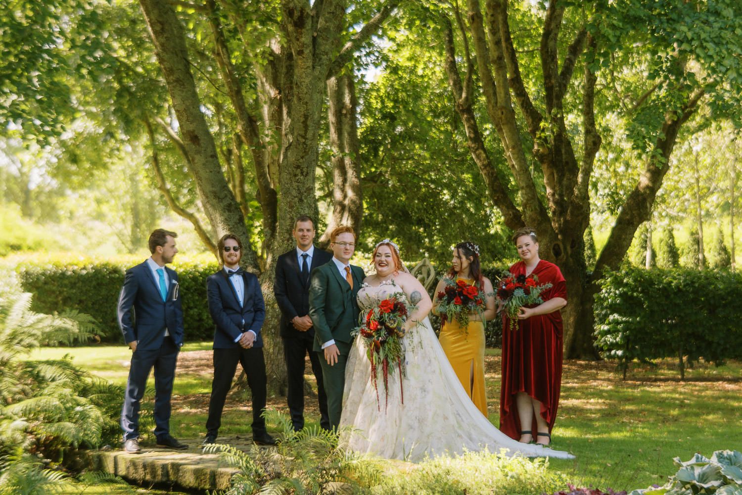 Wedding party poses outdoors under lush green trees, bride in white gown holding colorful bouquet with groomsmen and bridesmaids.