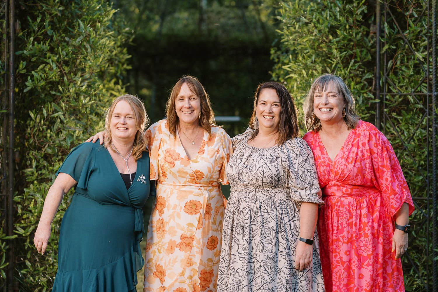 Four women in colorful dresses smiling together outdoors near lush green hedges at a garden event.