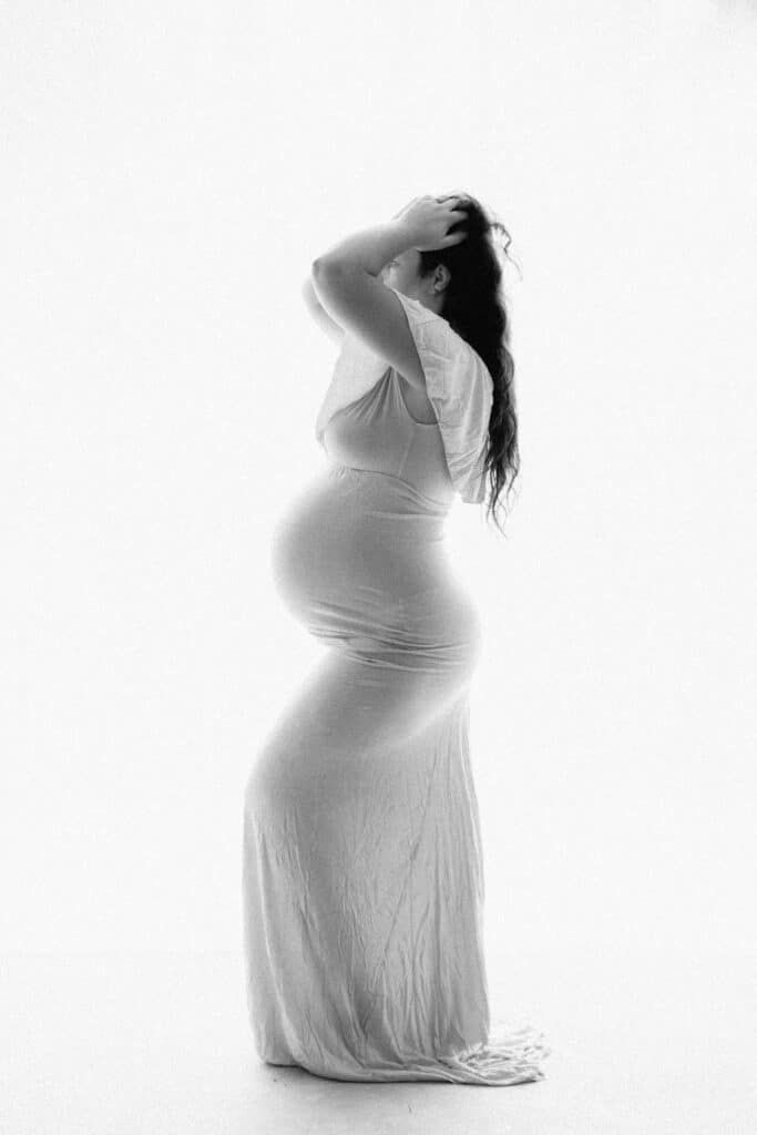 Black and white maternity photo of pregnant woman in white dress, side profile, hands in hair, studio background.