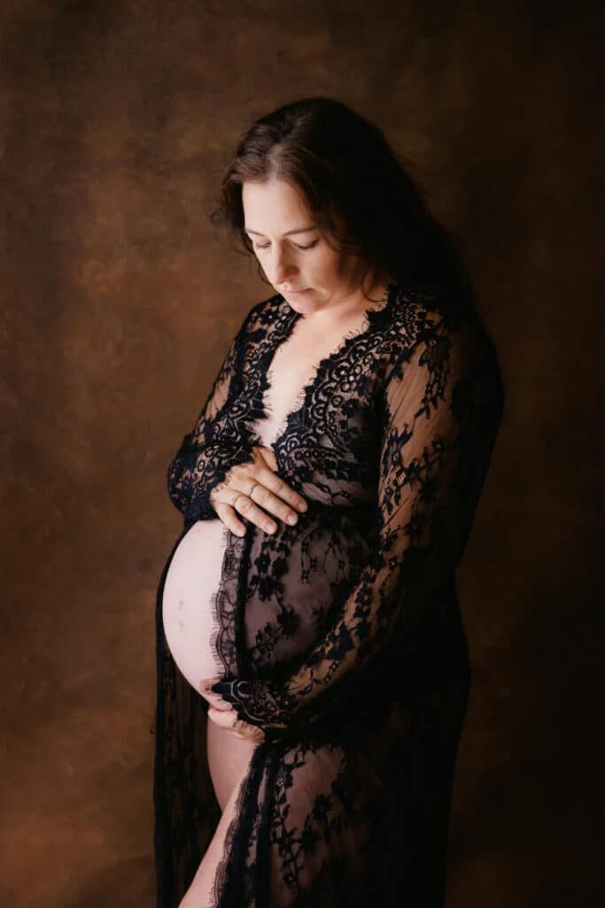 Pregnant woman in black lace maternity gown cradling baby bump in elegant studio portrait photography.