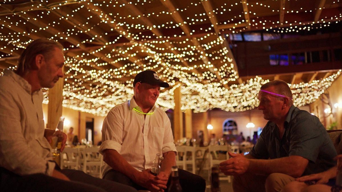 Three men socializing at an elegant event venue with warm string lights draped across the ceiling.