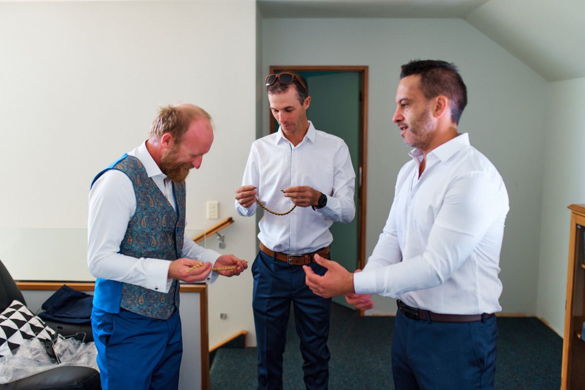 Three men in smart casual attire stand in a room, examining small items during what appears to be wedding preparations.