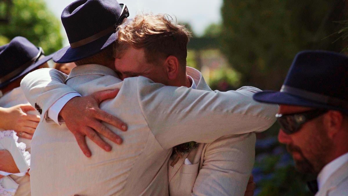 Men in suits and hats sharing a warm embrace at an outdoor wedding celebration.