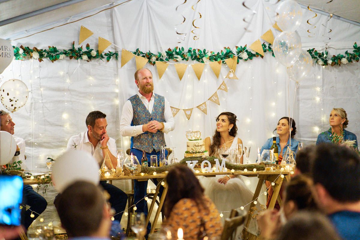 Best man gives a speech at a wedding reception in a decorated marquee with bunting and floral garlands.