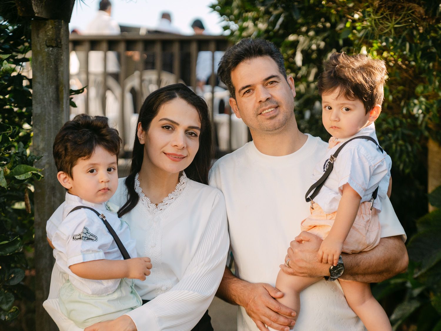 A happy family of four poses outdoors, parents holding two young boys dressed in white shirts with suspenders.