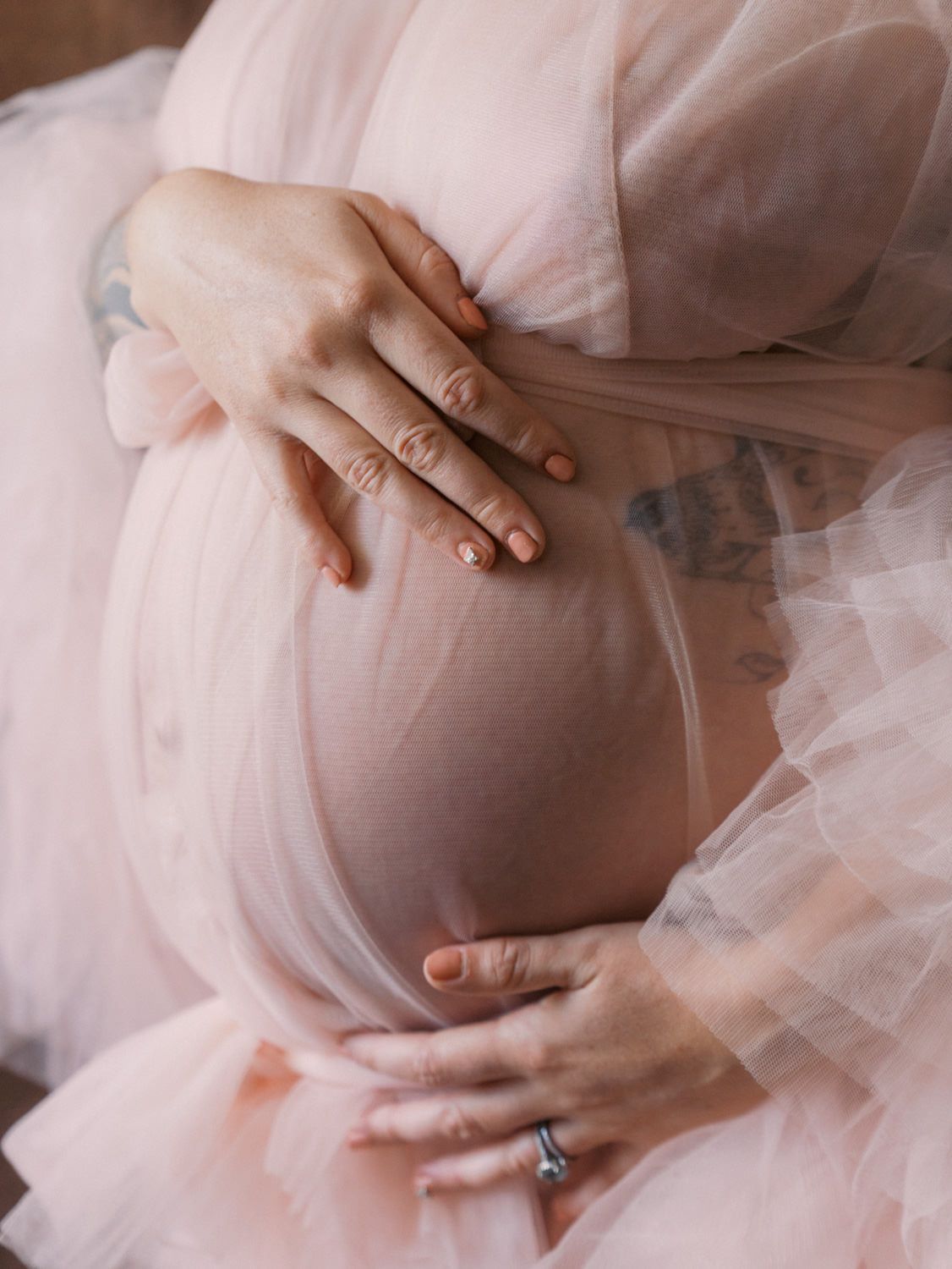 Pregnant woman in sheer pink tulle gown cradling bare baby bump with both hands, showing ring and tattoos.