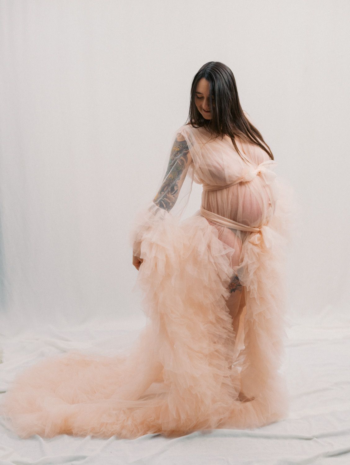 Pregnant woman in a flowing peach tulle maternity gown with tattoos, posing elegantly in a bright studio setting.