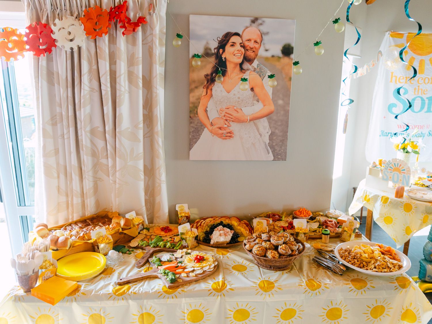 Festive baby shower food table with yellow daisy tablecloth, assorted dishes, and a wedding portrait backdrop.