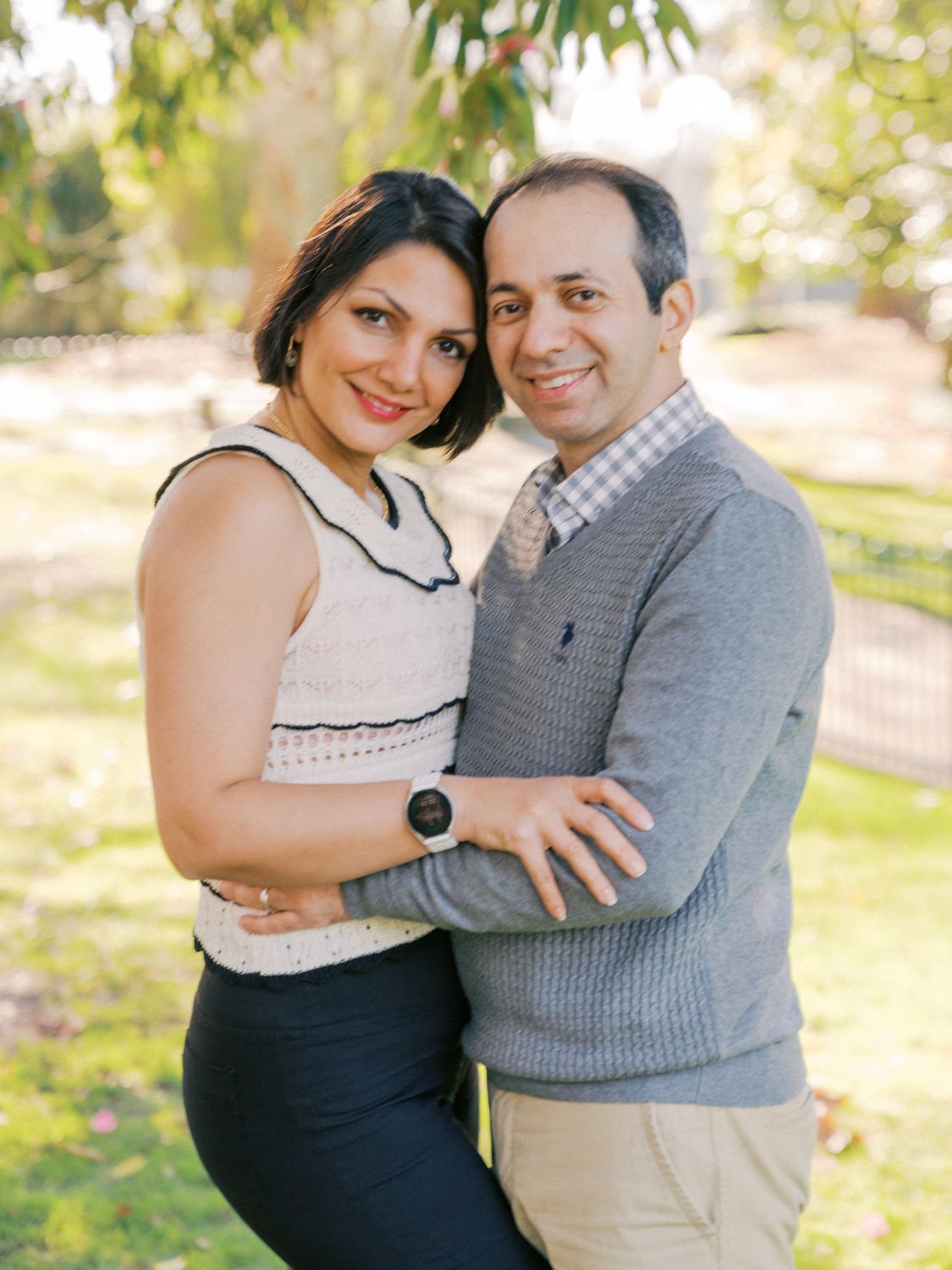 Smiling couple embracing outdoors in a park, woman in white top and man in gray sweater posing together.