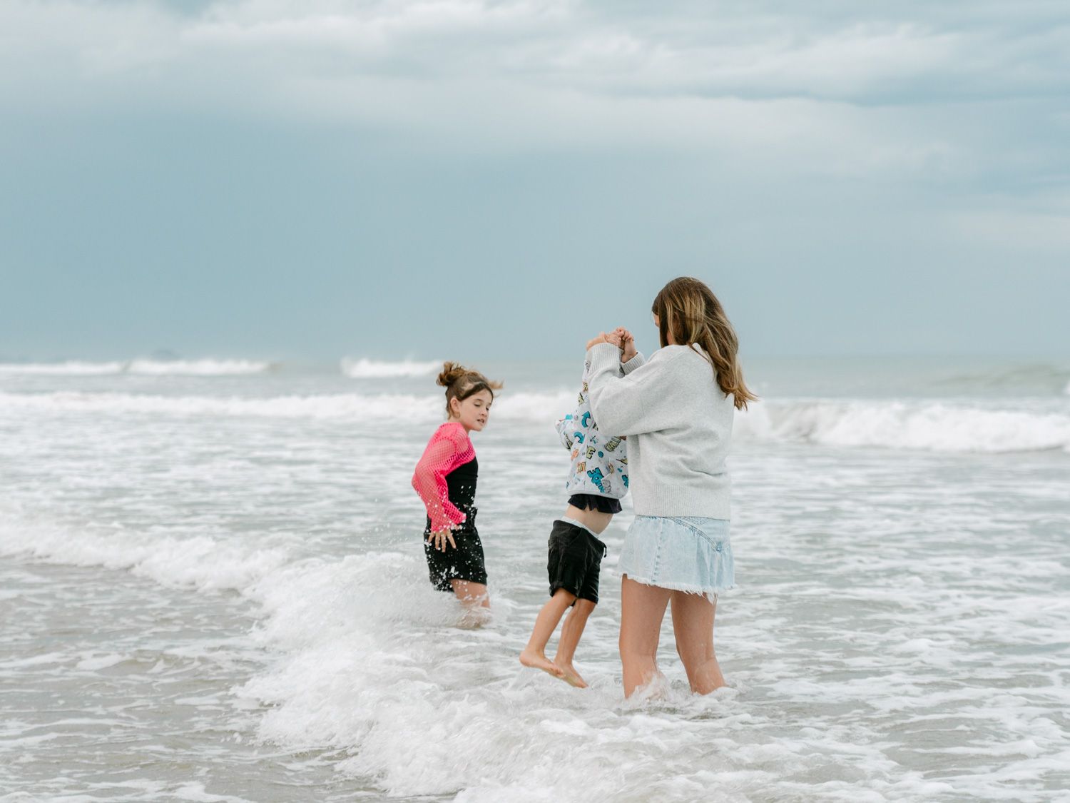 Two children and a woman playing in ocean waves on a sandy beach on a cloudy day.