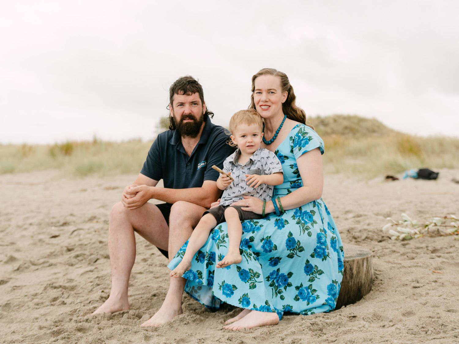 Family of three sitting on sandy beach, parents smiling with toddler in matching blue floral outfits on overcast day.