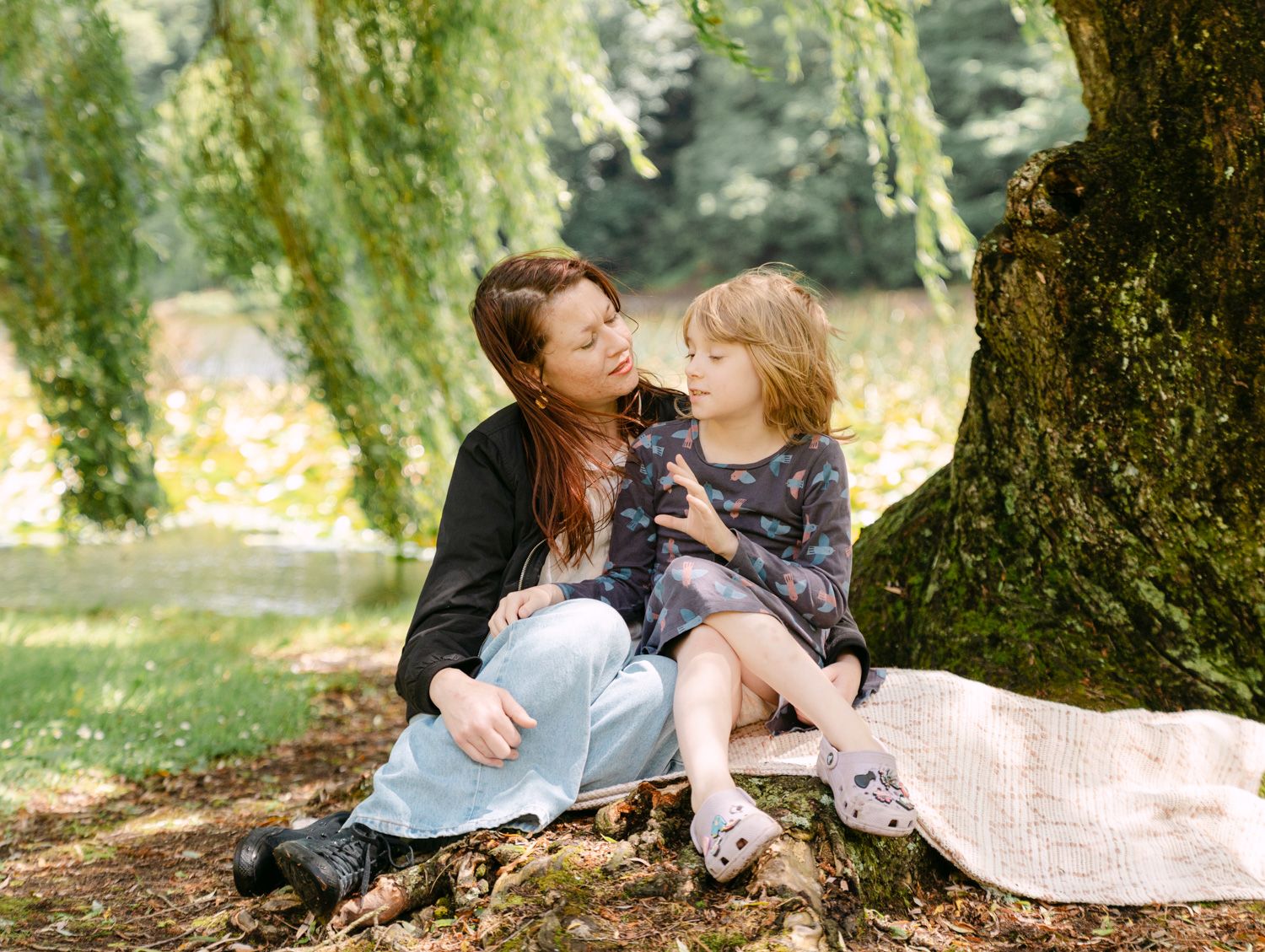 A mother and young daughter share a tender moment sitting under a weeping willow tree in a lush green park.