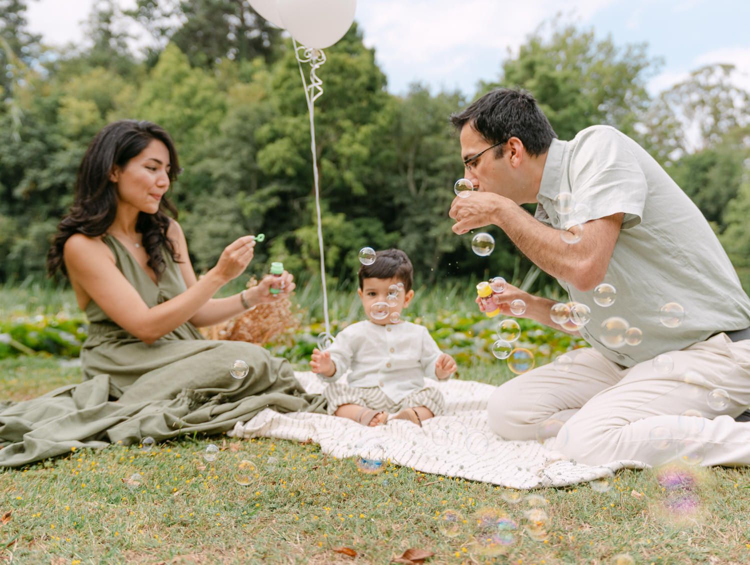 Family blowing bubbles with toddler on picnic blanket in lush green park on sunny day.