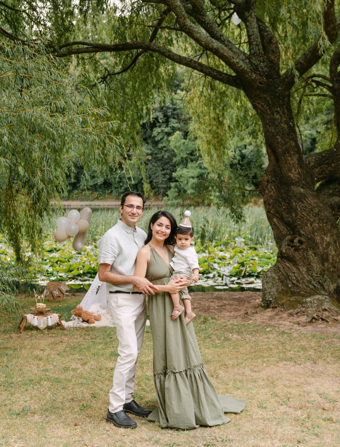 Happy family of three standing under a willow tree in a lush green garden, mother in sage green maxi dress.