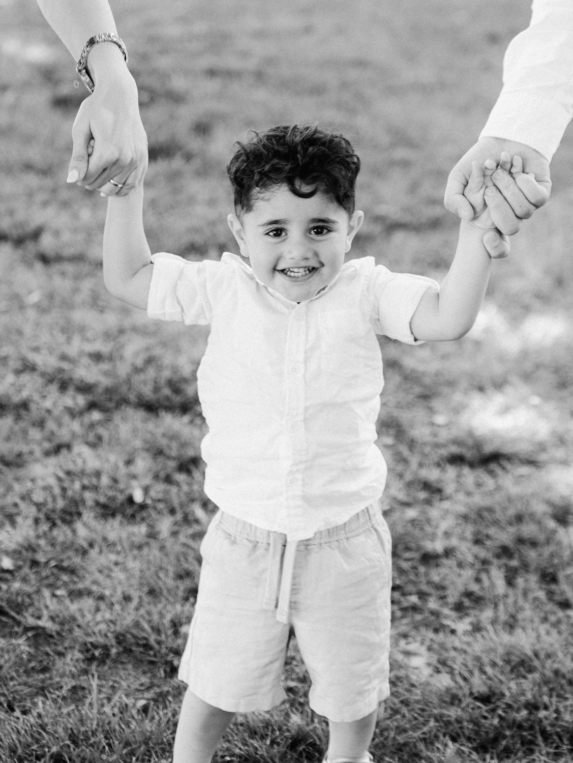 Black and white photo of a smiling young boy in white shirt and shorts holding hands with adults outdoors.