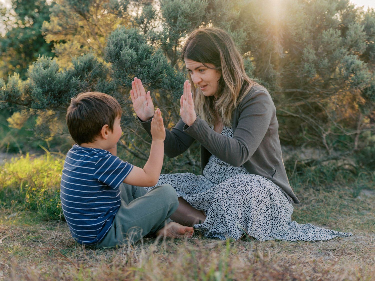 A mother and young boy playing hand games together while sitting outdoors among green trees.
