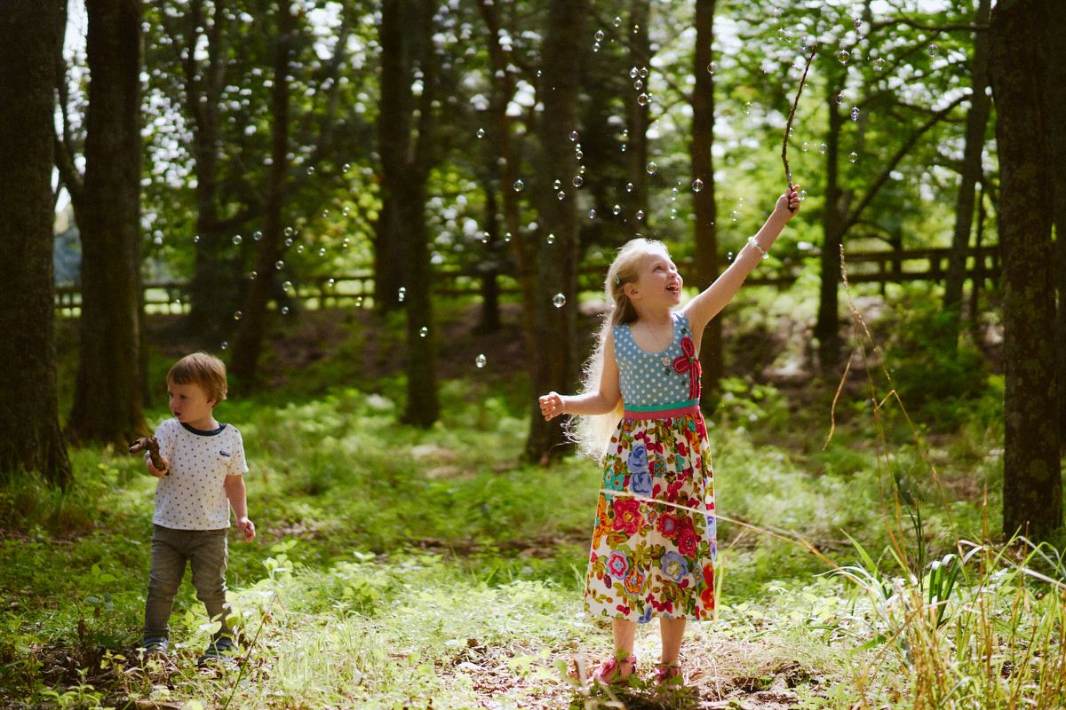 Two children playing with bubbles in a sunlit forest clearing surrounded by tall green trees.