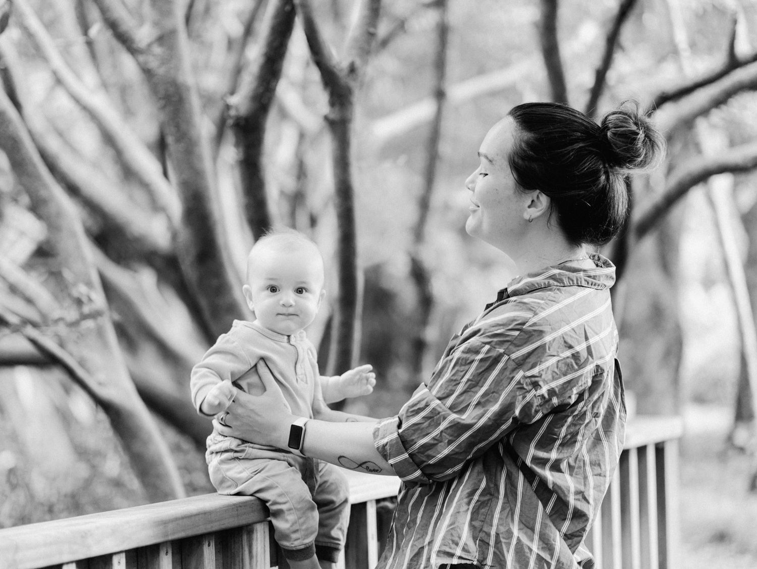 Black and white photo of a mother holding her baby on a wooden bridge surrounded by trees.