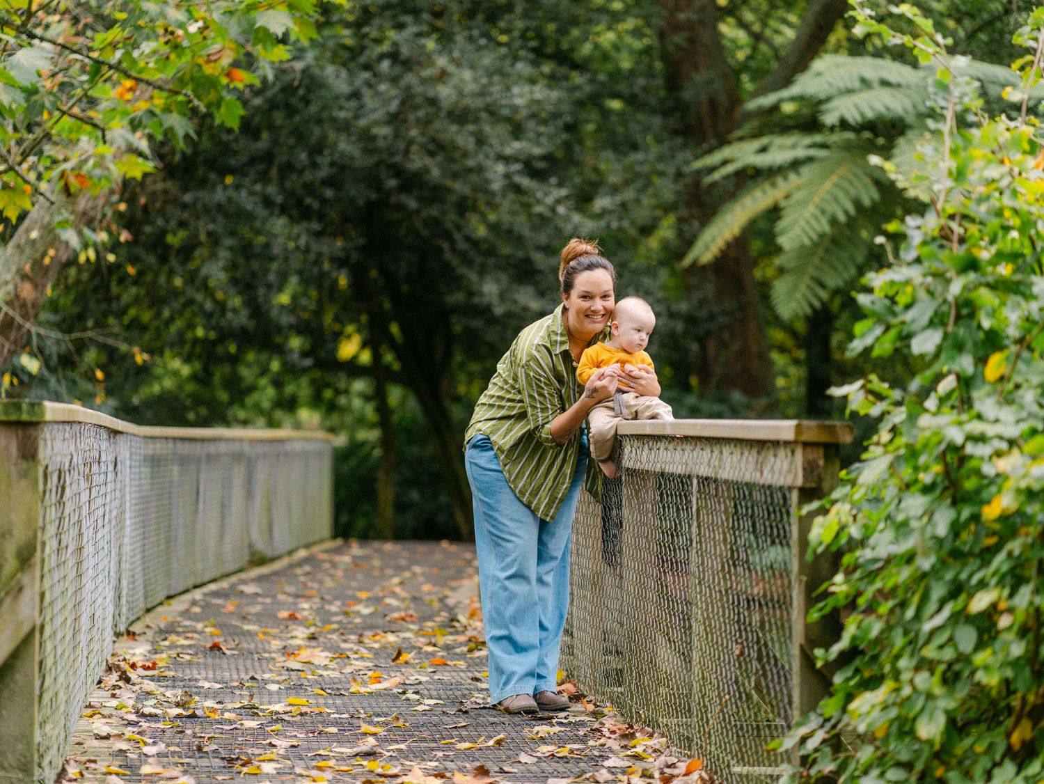 Mother holding baby on a leafy wooden bridge path surrounded by lush green trees in autumn.