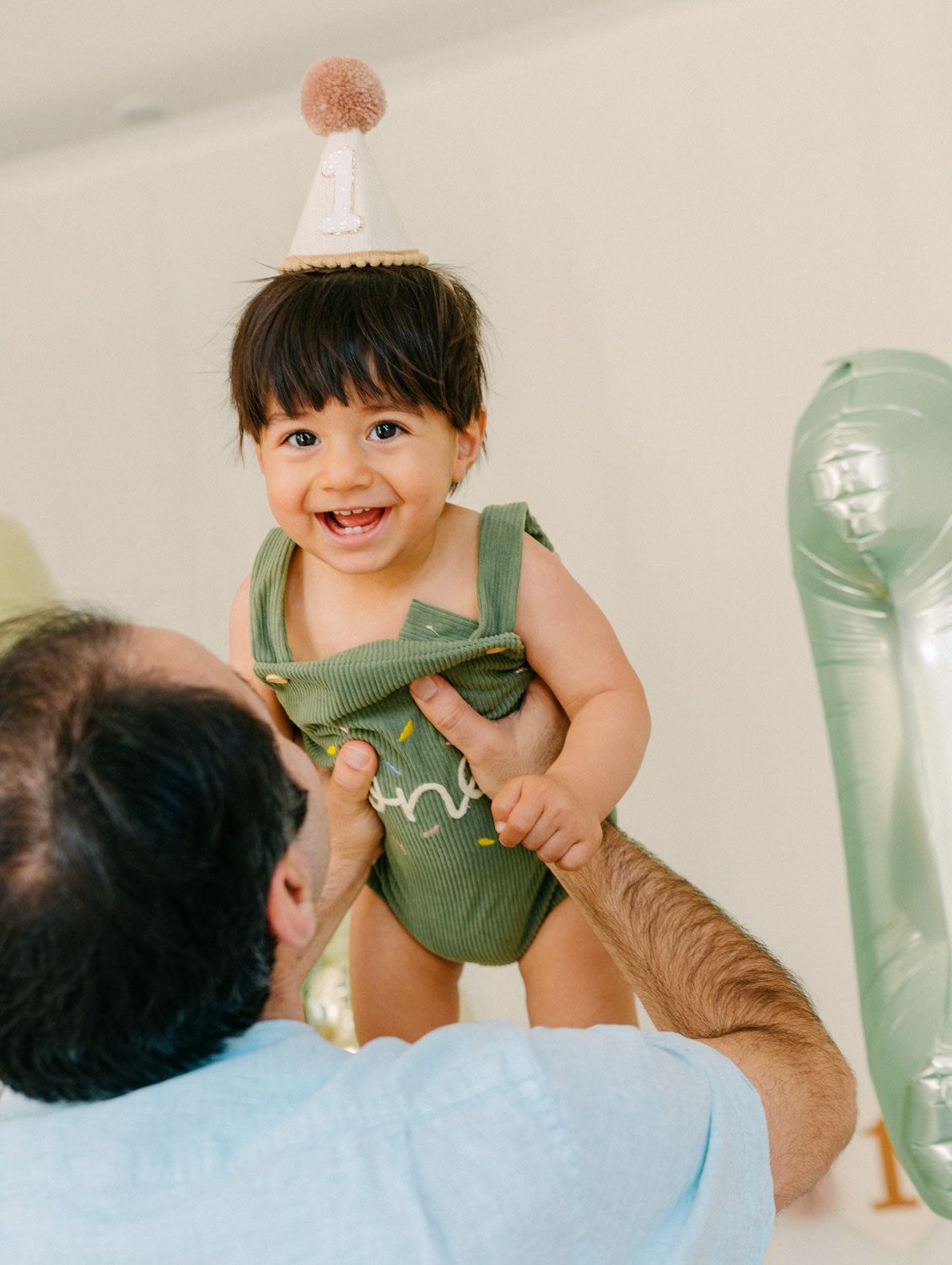 Happy baby in green romper and party hat being lifted by father at first birthday party.