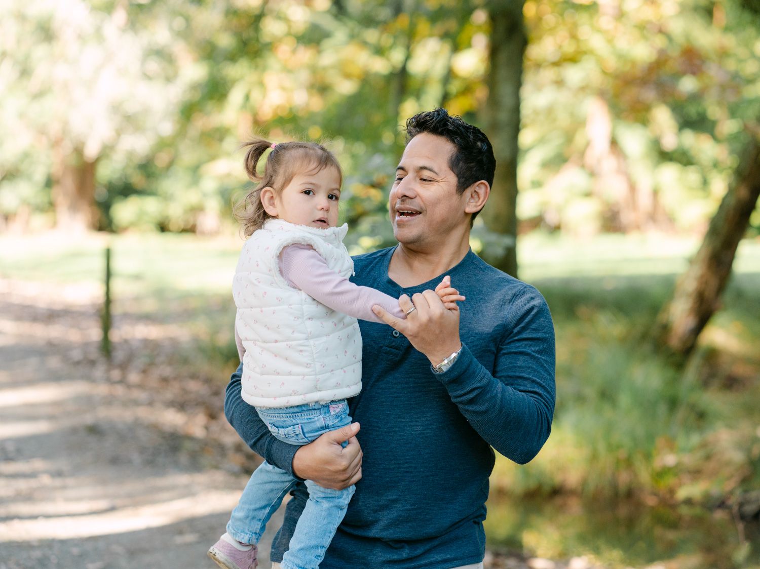 A man holding a toddler girl in a park, smiling and gesturing on a tree-lined path.