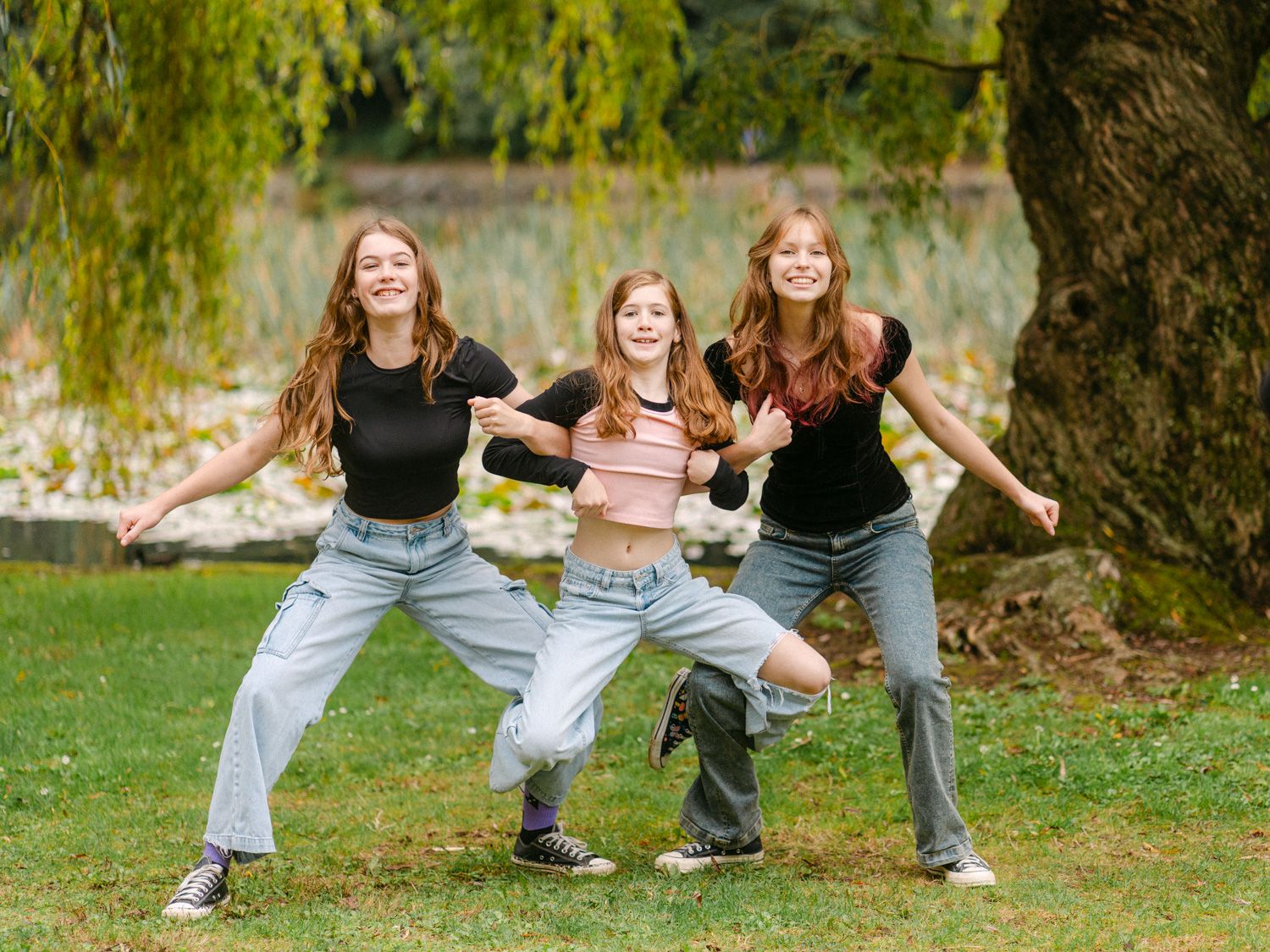 Three teenage girls dancing and posing playfully in a park with a large tree and green foliage backdrop.