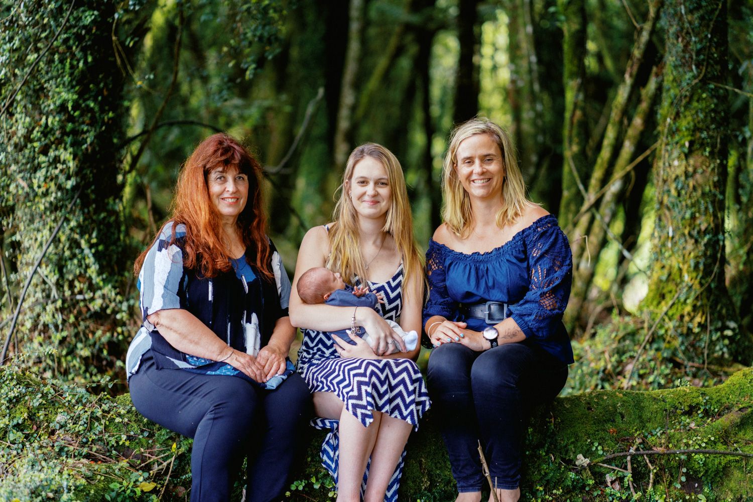 Three women sitting on a mossy log in a lush green forest, smiling together for a portrait photo.