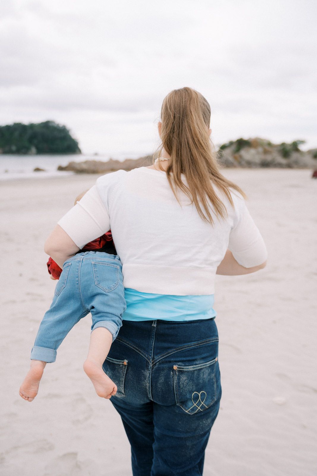 A woman carrying a young child on her hip while walking on a sandy beach, both seen from behind.