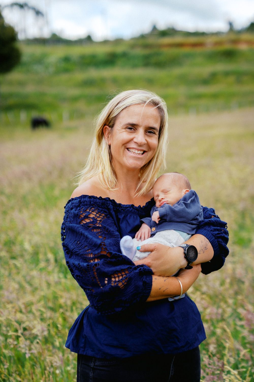 Smiling blonde woman in navy off-shoulder top holding sleeping newborn baby outdoors in green field.
