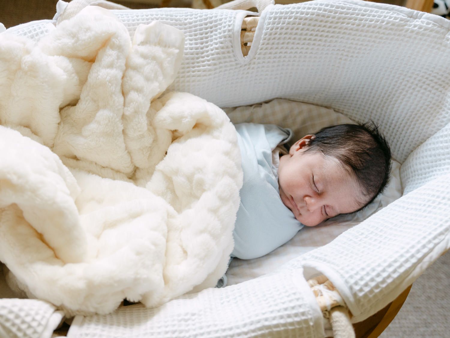 Sleeping newborn baby swaddled in light blue nestled in a wicker Moses basket with cream blankets.