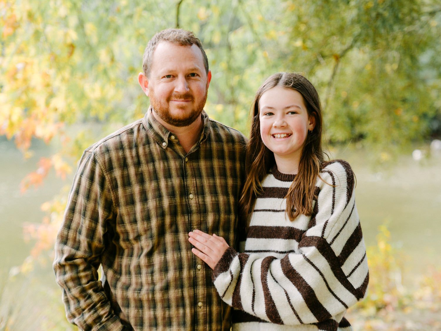 A man and young girl smile together outdoors in autumn, wearing casual clothes with colorful foliage in the background.