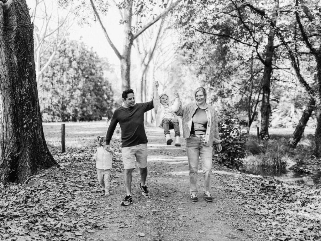 Black and white photo of a happy family walking on a park path, parents swinging child between them.