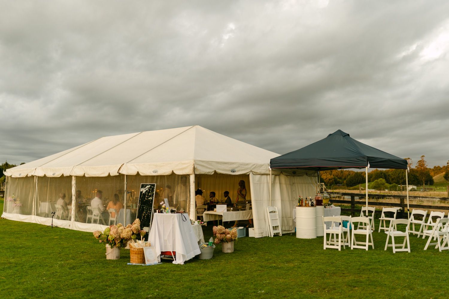 Outdoor wedding reception tent setup on green lawn with white chairs and floral arrangements under cloudy sky.