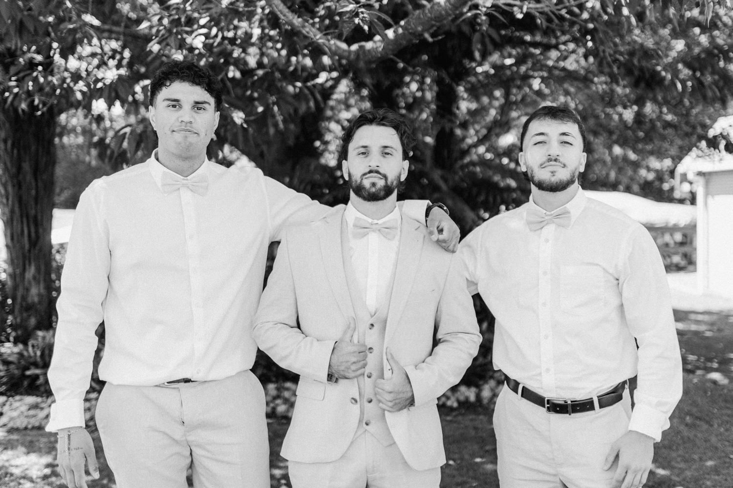 Three groomsmen in light suits and bow ties pose outdoors under trees in a classic black and white wedding photo.