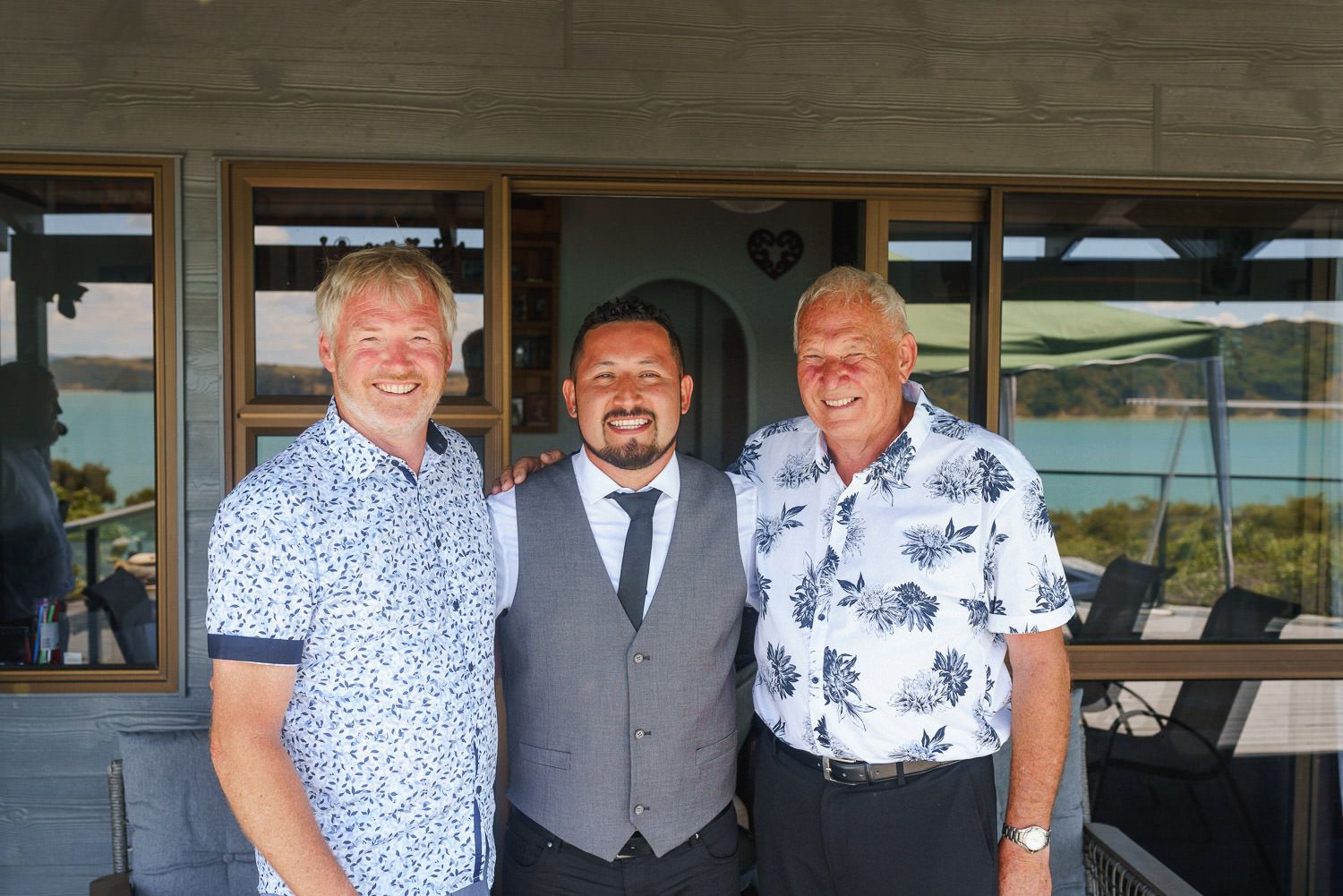 Three men smiling outdoors at a waterfront venue, one in a suit with tie flanked by two in casual shirts.