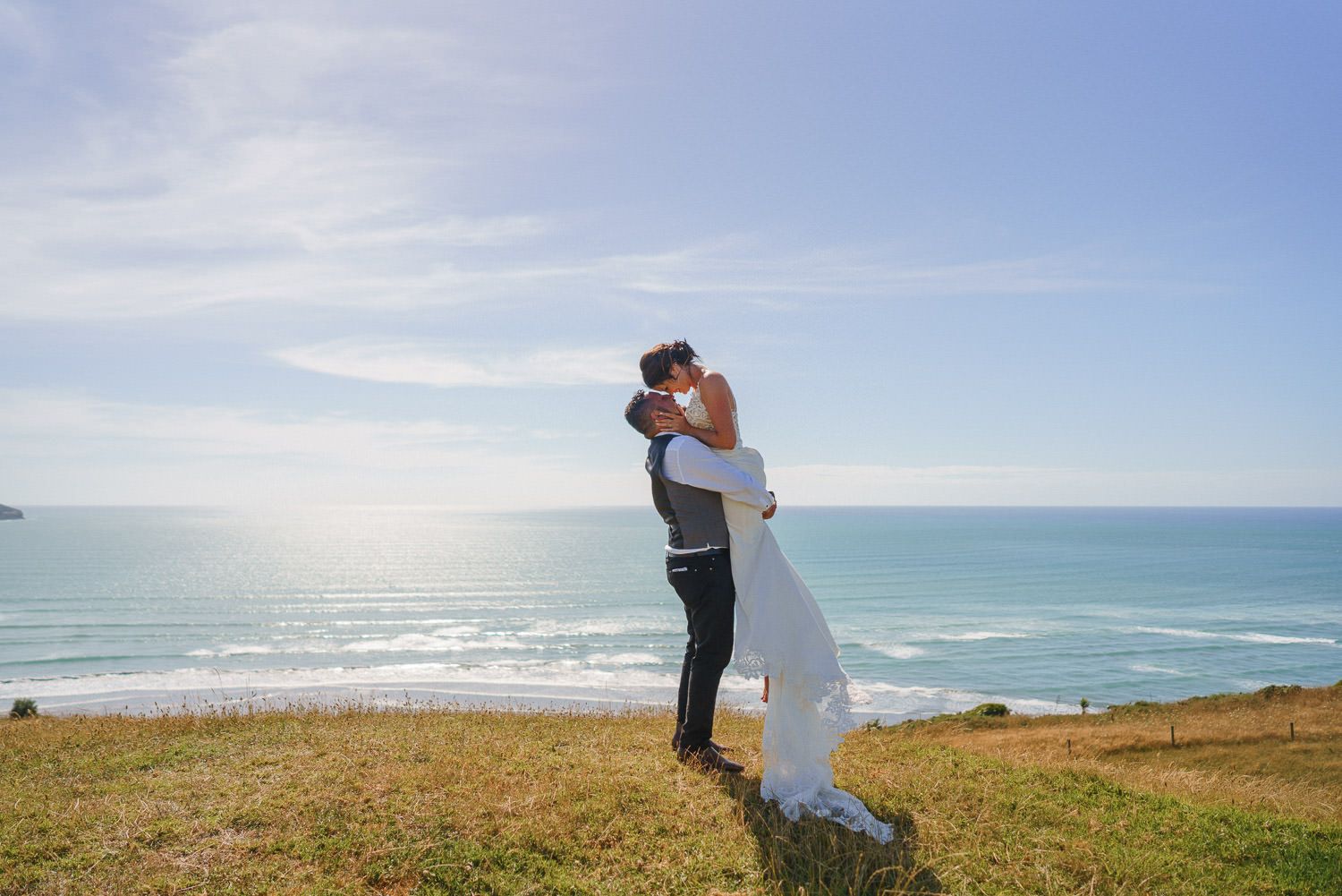 Bride and groom share romantic kiss on coastal cliff with turquoise ocean backdrop on sunny wedding day.
