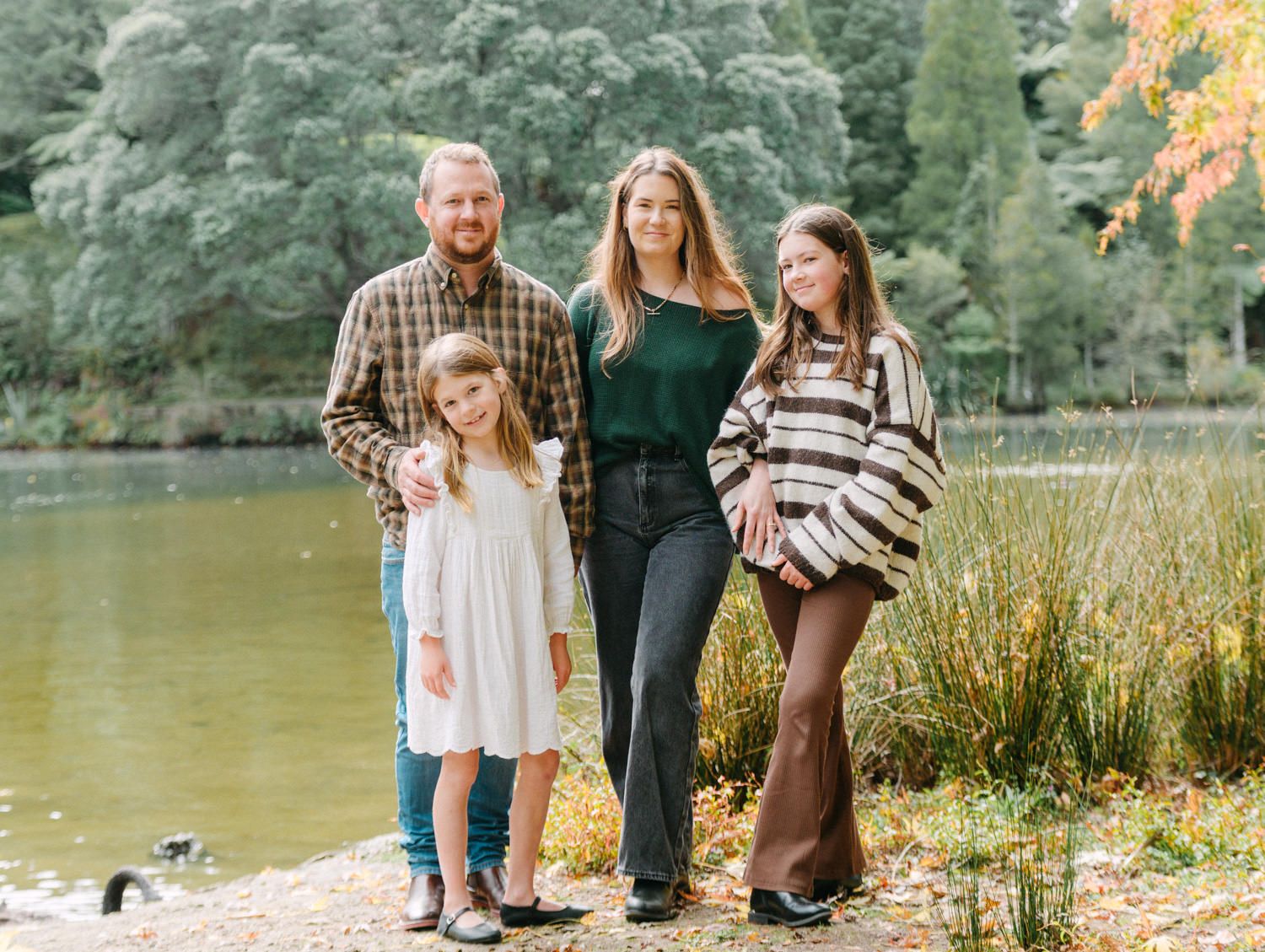 Family of four smiling outdoors near a pond surrounded by autumn foliage.
