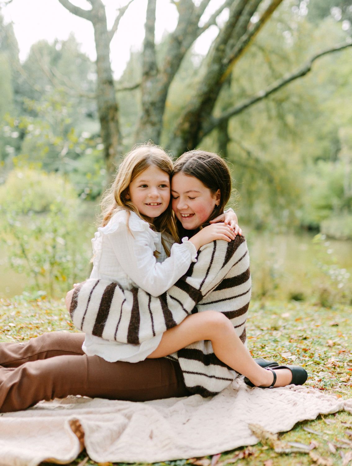 Two girls hugging and smiling on a blanket outdoors, surrounded by lush green trees in a candid family portrait.