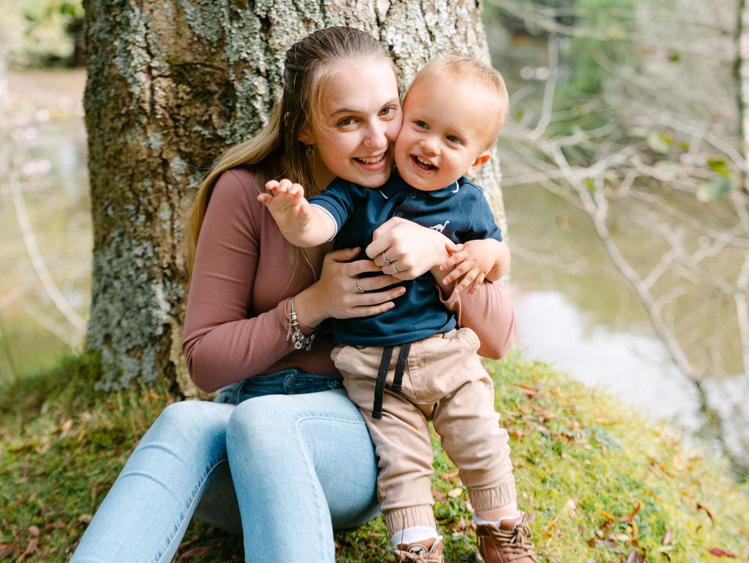 A smiling young mother sits against a tree holding her happy laughing baby boy outdoors.