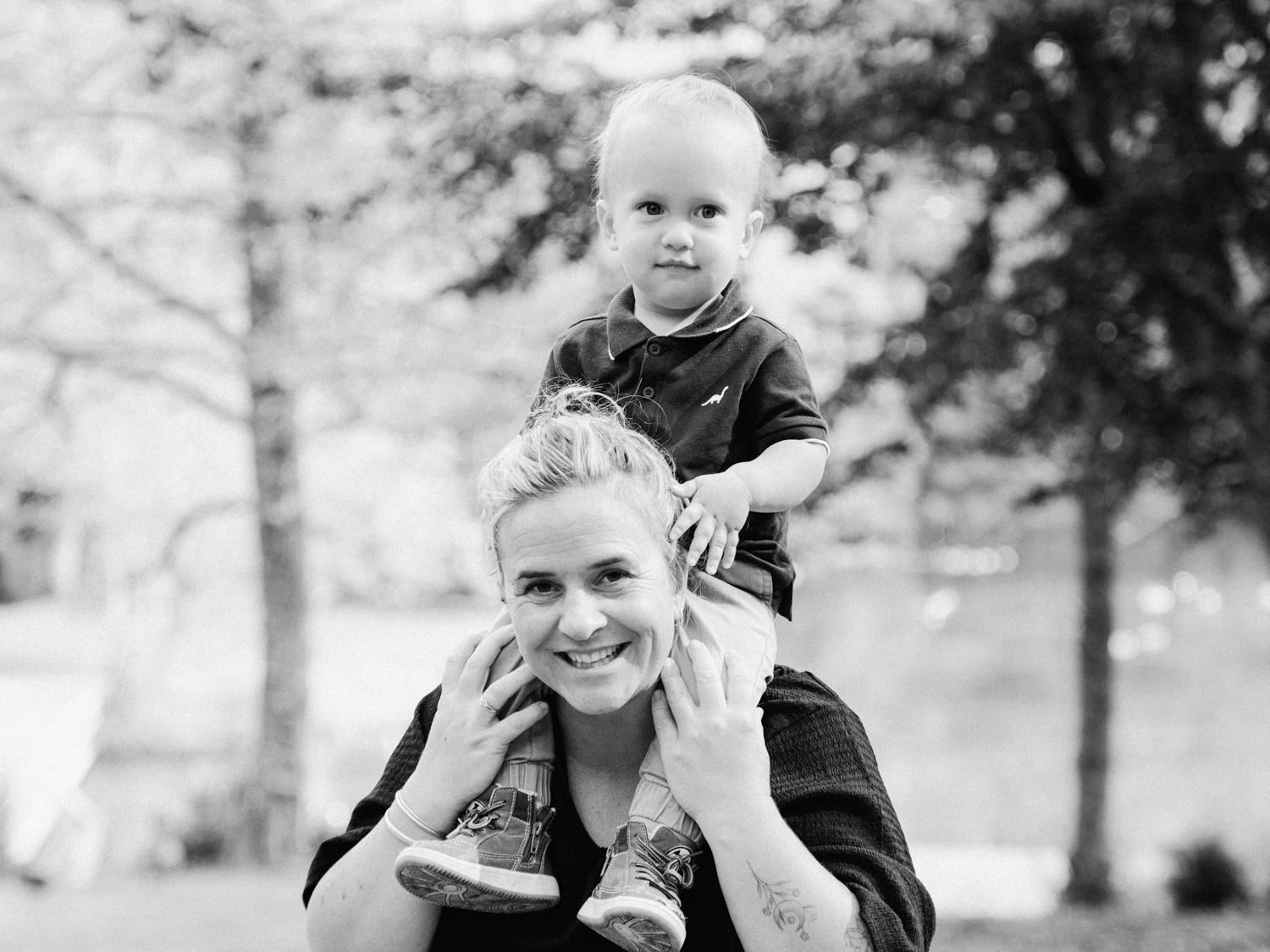Black and white photo of a happy mother with her toddler sitting on her shoulders outdoors.