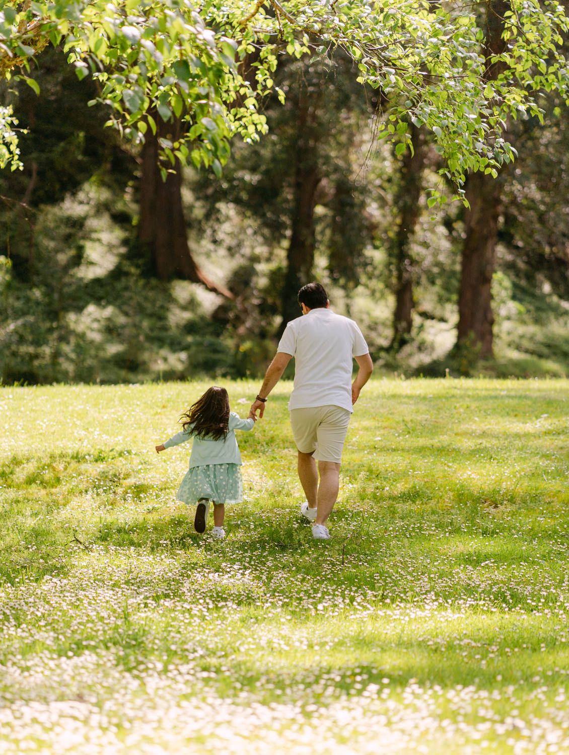 A father and young daughter run hand-in-hand through a sunlit park with lush green grass and trees.
