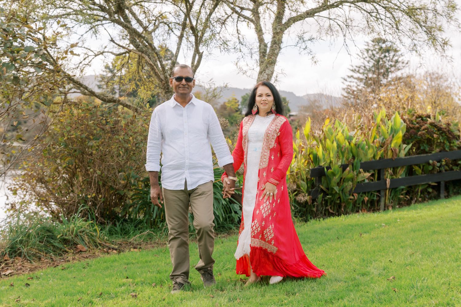 Couple in traditional South Asian attire holding hands on green lawn with trees and lake in background.